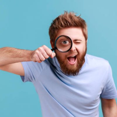 Portrait of funny positive bearded man holding magnifying glass and looking at camera with big zoom eye, curious face, keeps mouth open. Indoor studio shot isolated on blue background.